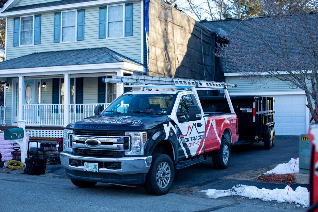 Work truck parked in front of a house with a black tarp on the roof and a trailer nearby.